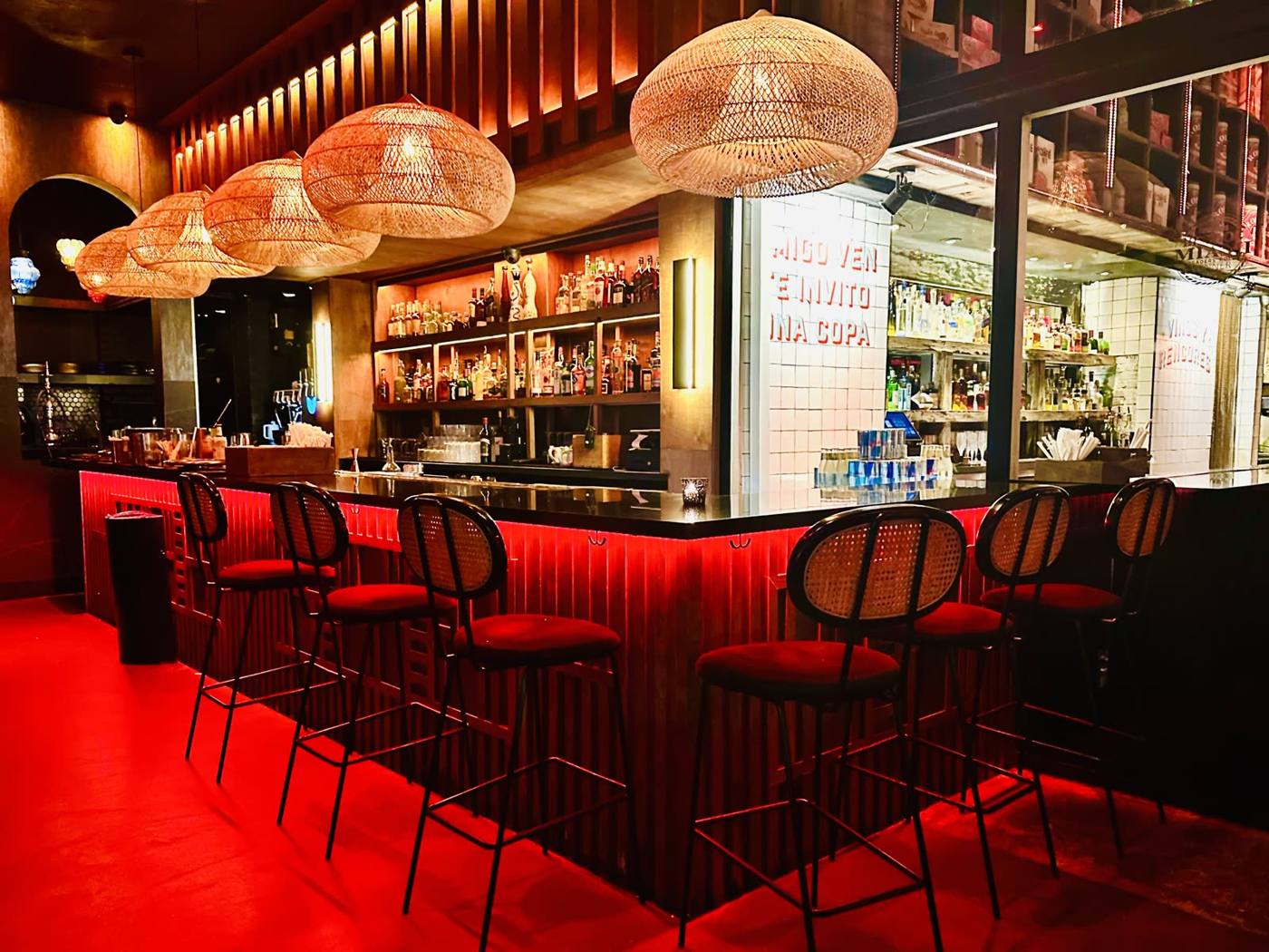 Bar interior with a long counter lit in red, wicker pendant lights overhead, and row of high stools in front of back-lit bottle shelves.