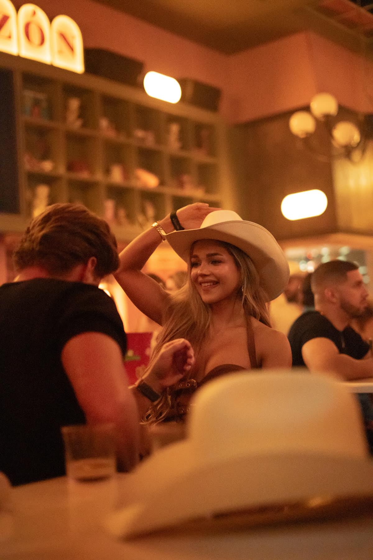 Smiling young woman wearing a wide-brimmed hat adjusts her hat in a warmly lit bar or nightclub setting.