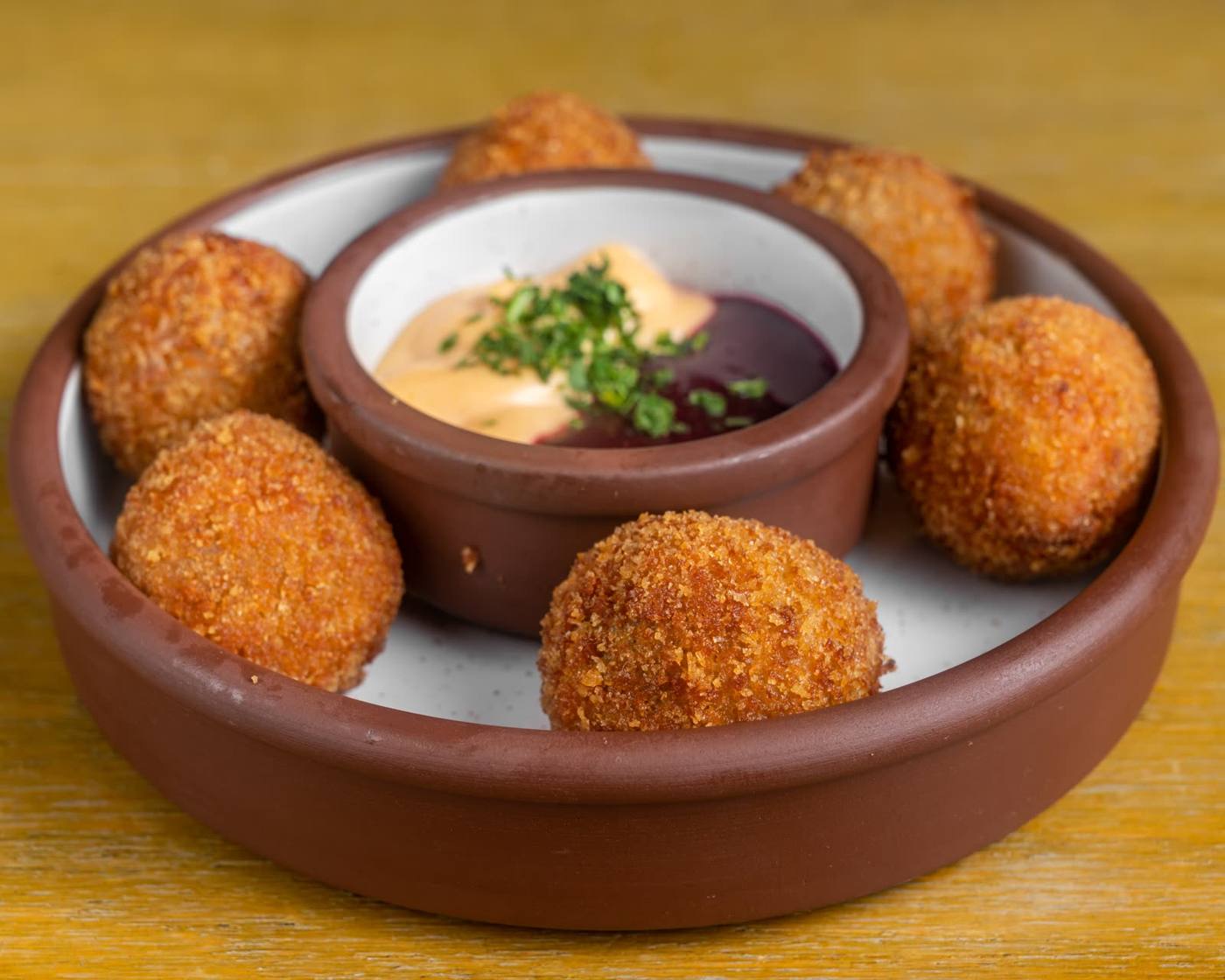 Round clay plate with golden-brown croquettes surrounding a dipping sauce duo (creamy orange and dark red) garnished with chopped herbs in the center dip cup.