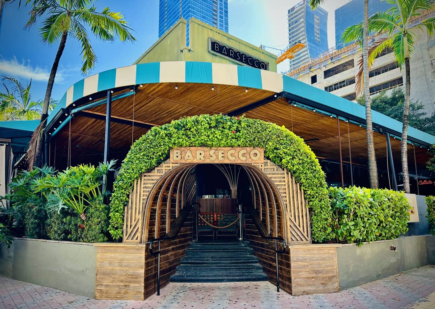 Entrance to Barsecco restaurant with a curved wooden archway framed by lush ivy and steps leading inside, tropical plants around.