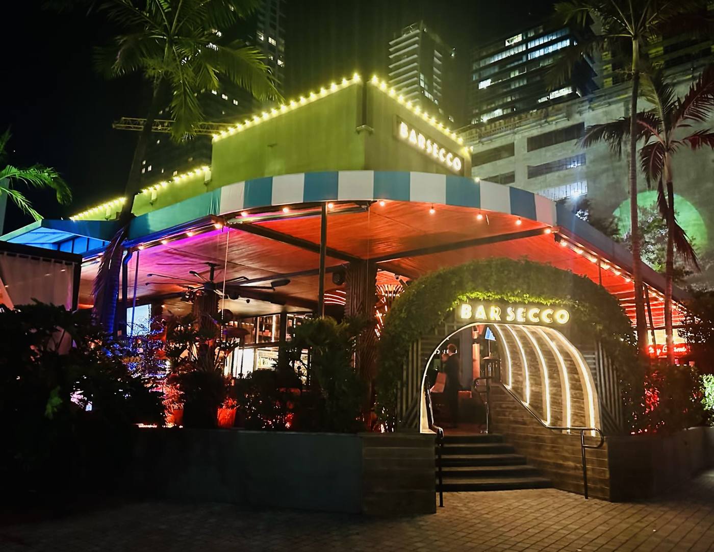 Nighttime exterior of Barsecco with colorful lights, striped canopy, and a curved, illuminated entrance arch.