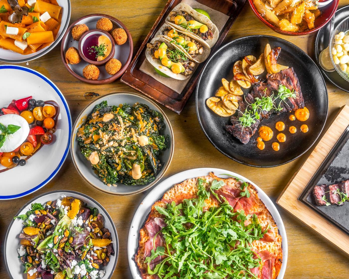Colorful assortment of dishes on a wooden table: pizza, greens, pasta, tacos, roasted meat, and roasted potatoes for a shared meal.