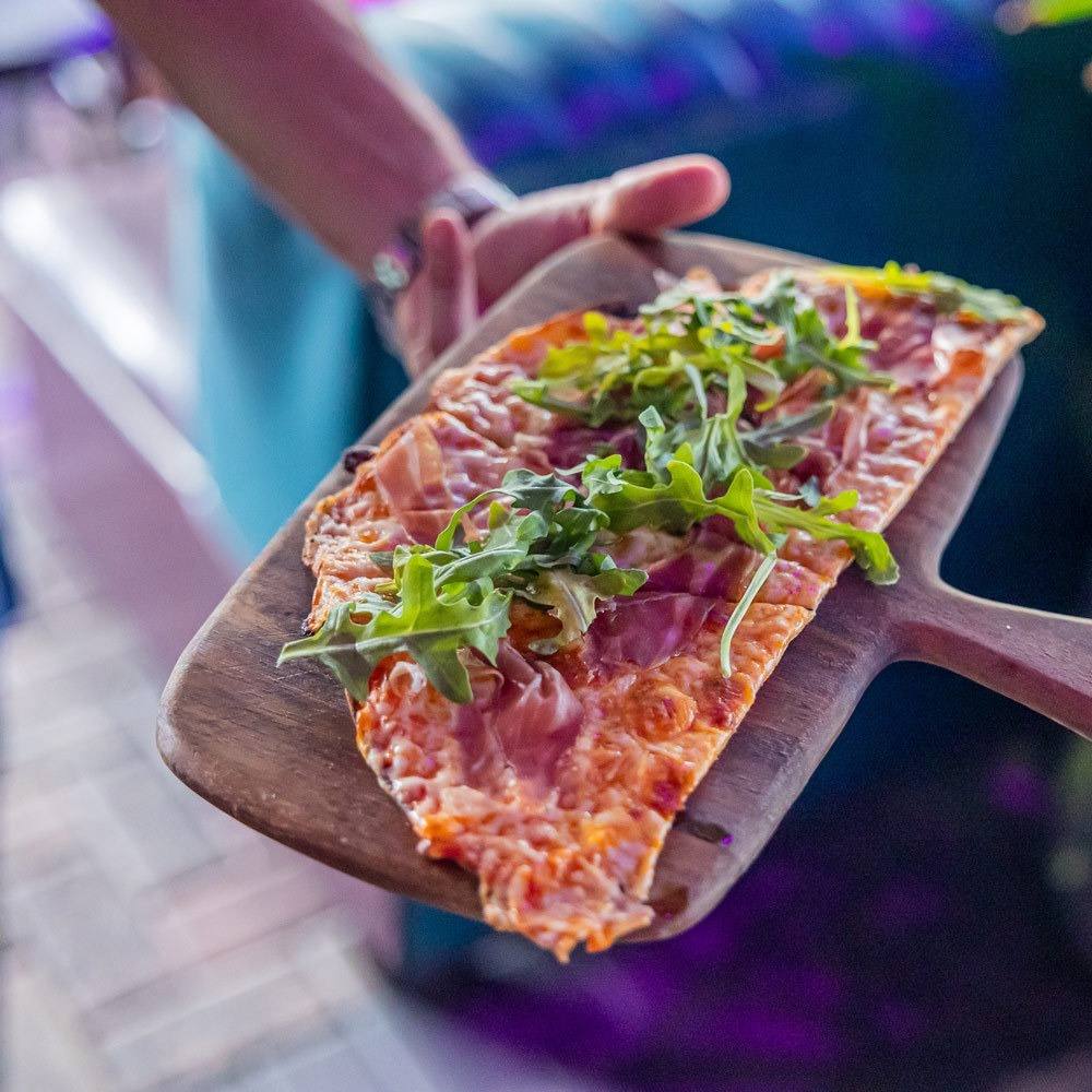 Flat rectangular flatbread pizza topped with greens on a wooden paddle being held by a person's hand