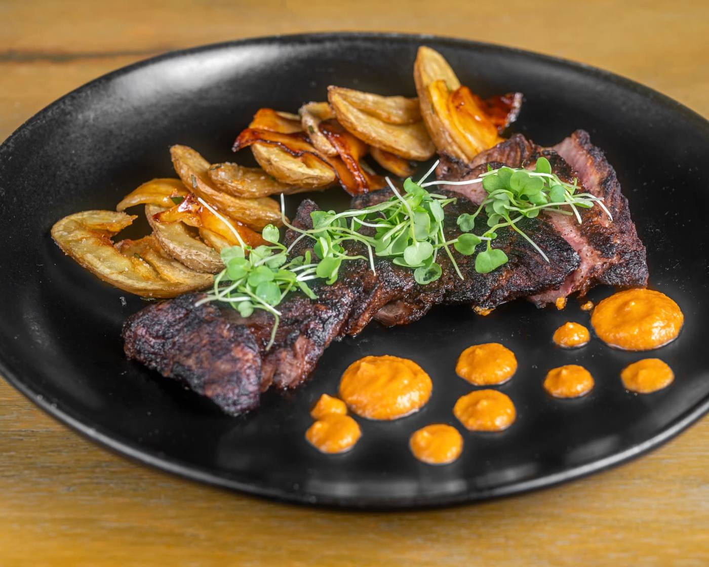 Barbecue ribs with microgreen garnish, crispy potato wedges, and orange sauce dots on a black plate.