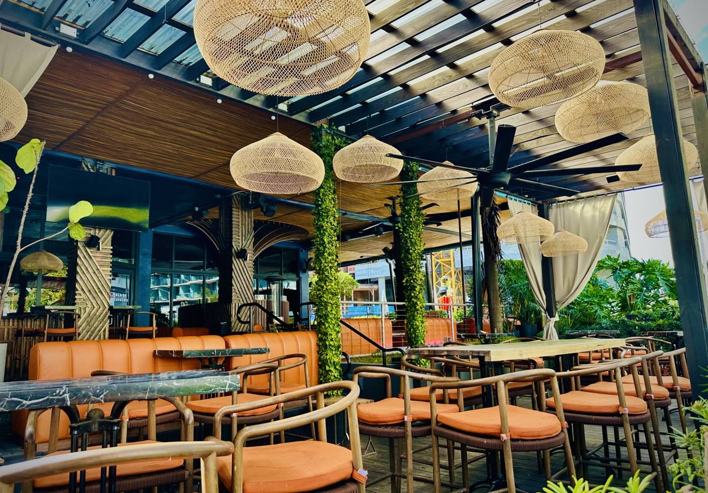 Outdoor cafe with orange cushioned chairs and marble-topped tables under a glass ceiling with woven wicker pendant lights and greenery.